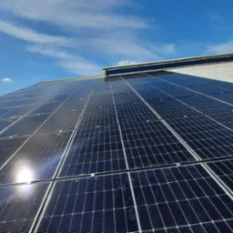 Instalacja paneli fotowoltaicznych na metalowym dachu budynku przemysłowego, widok pod kątem z odbiciem słońca na panelach i błękitnym niebem z chmurami w tle.