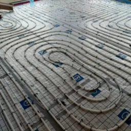 Instalacja ogrzewania podłogowego z rur ułożonych w spiralny wzór na izolacji podłogowej w surowym stanie, widoczne okno i butelki z wodą.