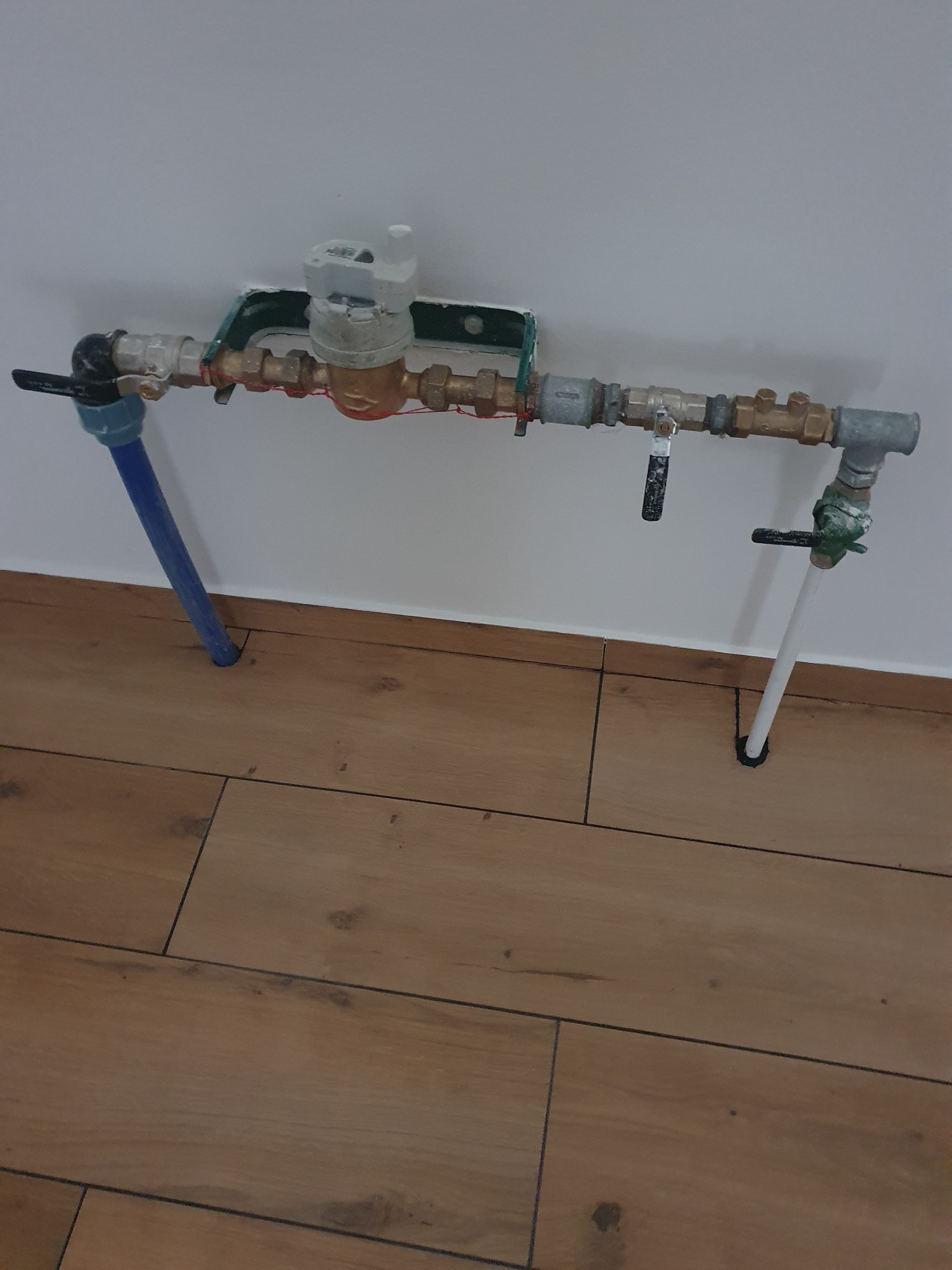 Instalacja wodociągowa z licznikiem, rurami miedzianymi i zaworami, przymocowana do ściany nad drewnianą podłogą.