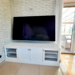 Biała szafka RTV z przeszklonymi półkami po bokach, stojąca pod dużym telewizorem na tle ściany z białej cegły i okna z białymi zasłonami.