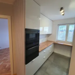 Nowoczesna kuchnia z białymi, lakierowanymi szafkami, drewnianym blatem i zabudowanym sprzętem AGD (piekarnik i mikrofalówka) w czarnej obudowie, uzupełniona o dekoracyjny panel z drewnianych...