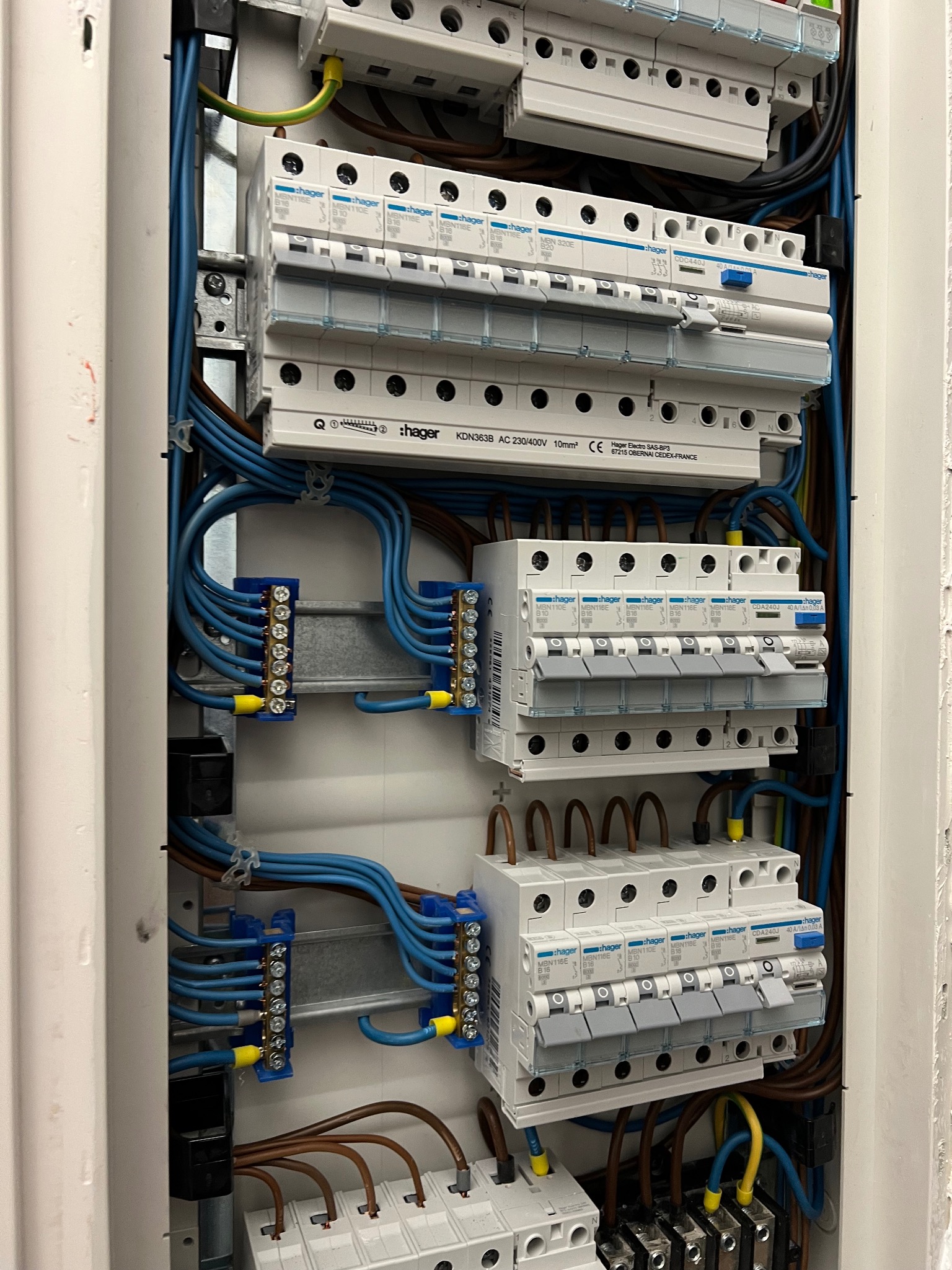 Rozdzielnica elektryczna Hager z niebieskimi i brązowymi przewodami, widoczne bezpieczniki i listwy zaciskowe. Instalacja w białej obudowie, detale z bliska.