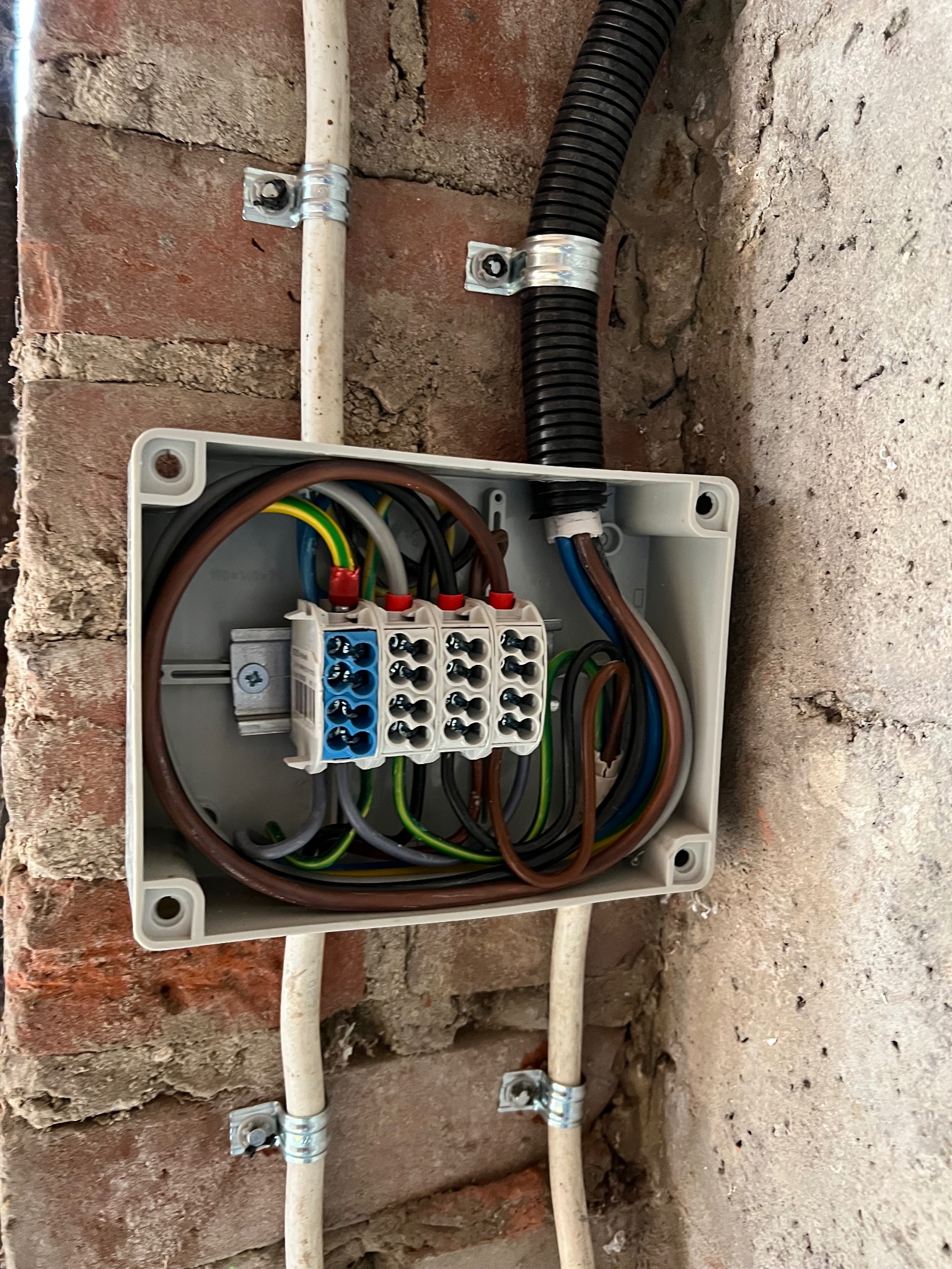 Otwarta puszka elektryczna z widocznymi przewodami i złączkami, zamontowana na surowej ścianie z cegły i betonu. Widoczne kable w peszlu i rurkach PCV.