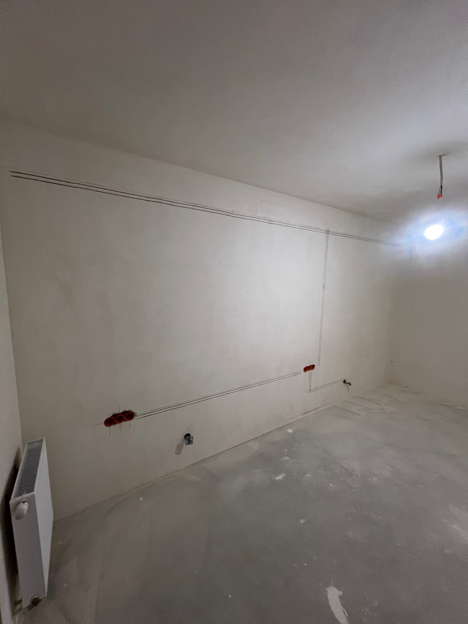 Ściana z nową instalacją elektryczną: kable w peszlach, puszki pod gniazdka. Widoczny grzejnik i surowa podłoga. Modernizacja instalacji po deweloperze.