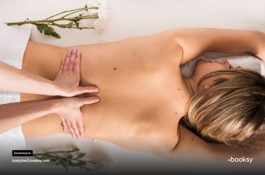 Kobieta leżąca na brzuchu podczas masażu pleców; dłonie masażysty uciskają dolną część pleców, w tle białe kwiaty i ręcznik.