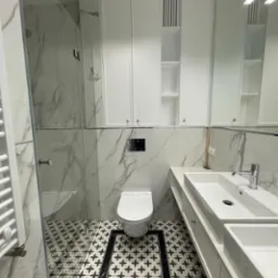 Nowoczesna łazienka z białymi szafkami, marmurowymi płytkami ściennymi i wzorzystą podłogą w czarno-białe kwiaty, z kabiną prysznicową ze szklaną ścianą i grzejnikiem drabinkowym.