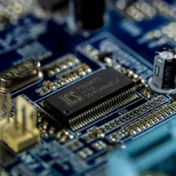 Zbliżenie na niebieską płytę główną komputera z widocznymi elementami elektronicznymi: procesorem ICS z numerem 7323873 0702 954148AELF, kondensatorem TEAPO i innymi komponentami. Płytka oświetlona...