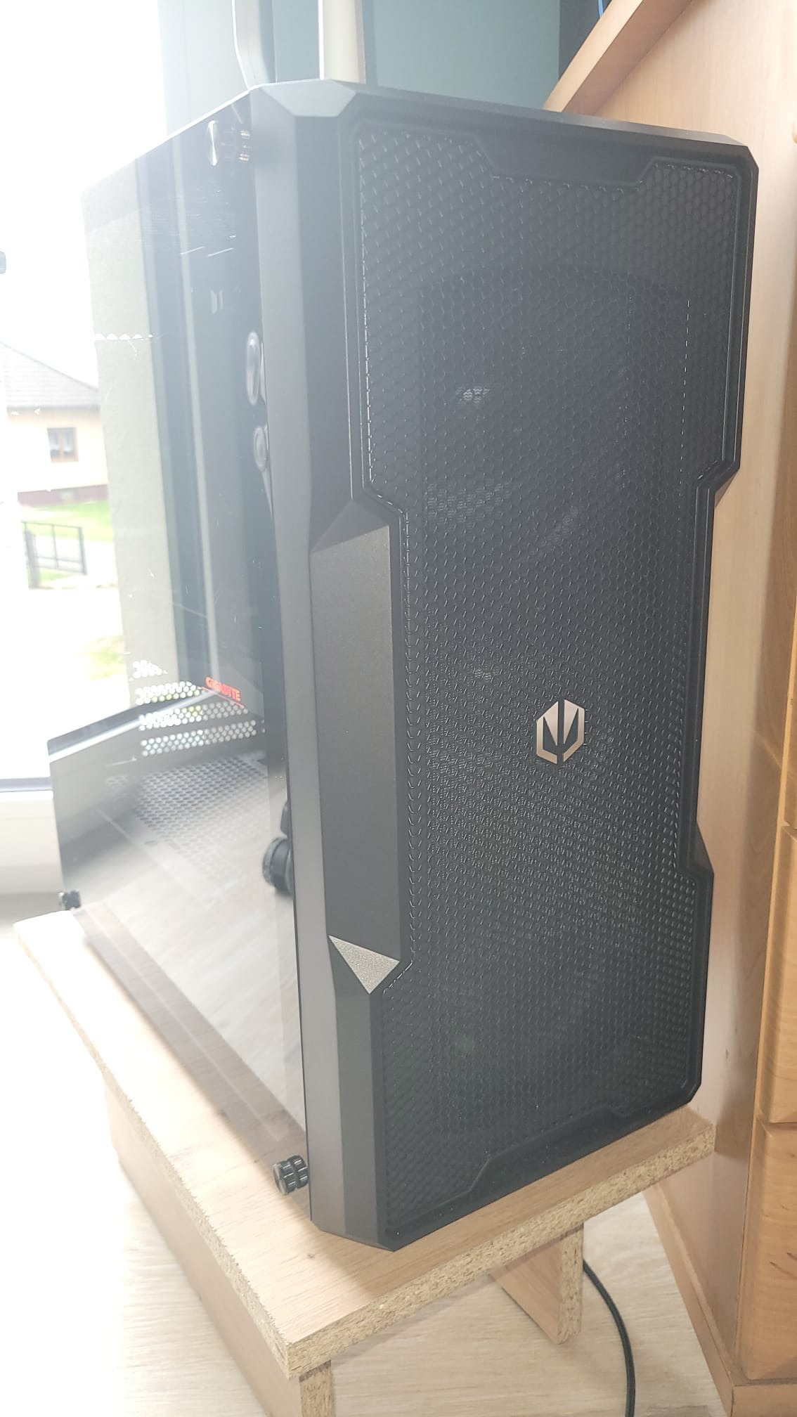 Ciemna obudowa komputera stacjonarnego z panelem bocznym z hartowanego szkła, umieszczona na drewnianym podeście, z widocznym logo producenta na perforowanym froncie.