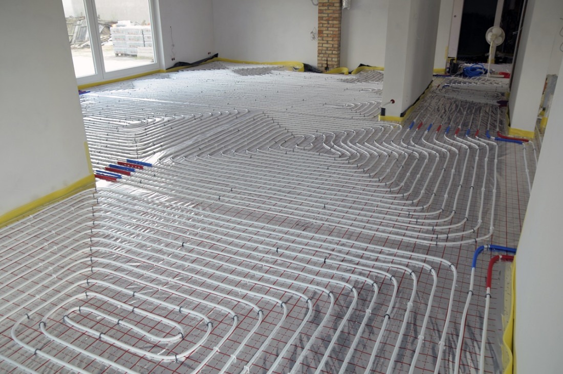 Rozłożone białe rury ogrzewania podłogowego na folii z czerwoną siatką w surowym wnętrzu, widoczne niebieskie i czerwone złączki przy ścianie, żółta taśma zabezpieczająca na obrzeżach podłogi.
