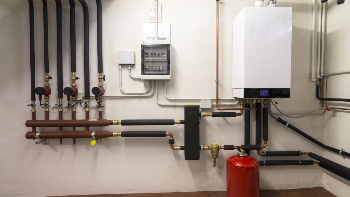 Nowoczesna instalacja centralnego ogrzewania z białym piecem, czerwonym zbiornikiem wyrównawczym, pompami obiegowymi i rozdzielnicą elektryczną na ścianie w kotłowni.