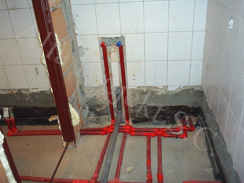 Łazienka w trakcie remontu: czerwone rury instalacji wodnej ułożone na podłodze, białe płytki na ścianach, widoczny fragment ościeżnicy z pianką montażową.