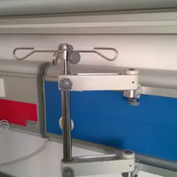 Metalowy, regulowany wieszak na kroplówki przymocowany do ściany w szpitalnej sali, z widocznymi elementami mocującymi i regulacyjnymi.