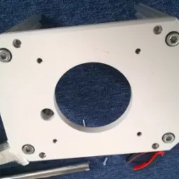 Biała, metalowa płyta z centralnym okrągłym otworem i kilkoma mniejszymi otworami na śruby, leżąca na niebieskiej, teksturowanej powierzchni, obok narzędzi takich jak kombinerki i fragmenty profili...