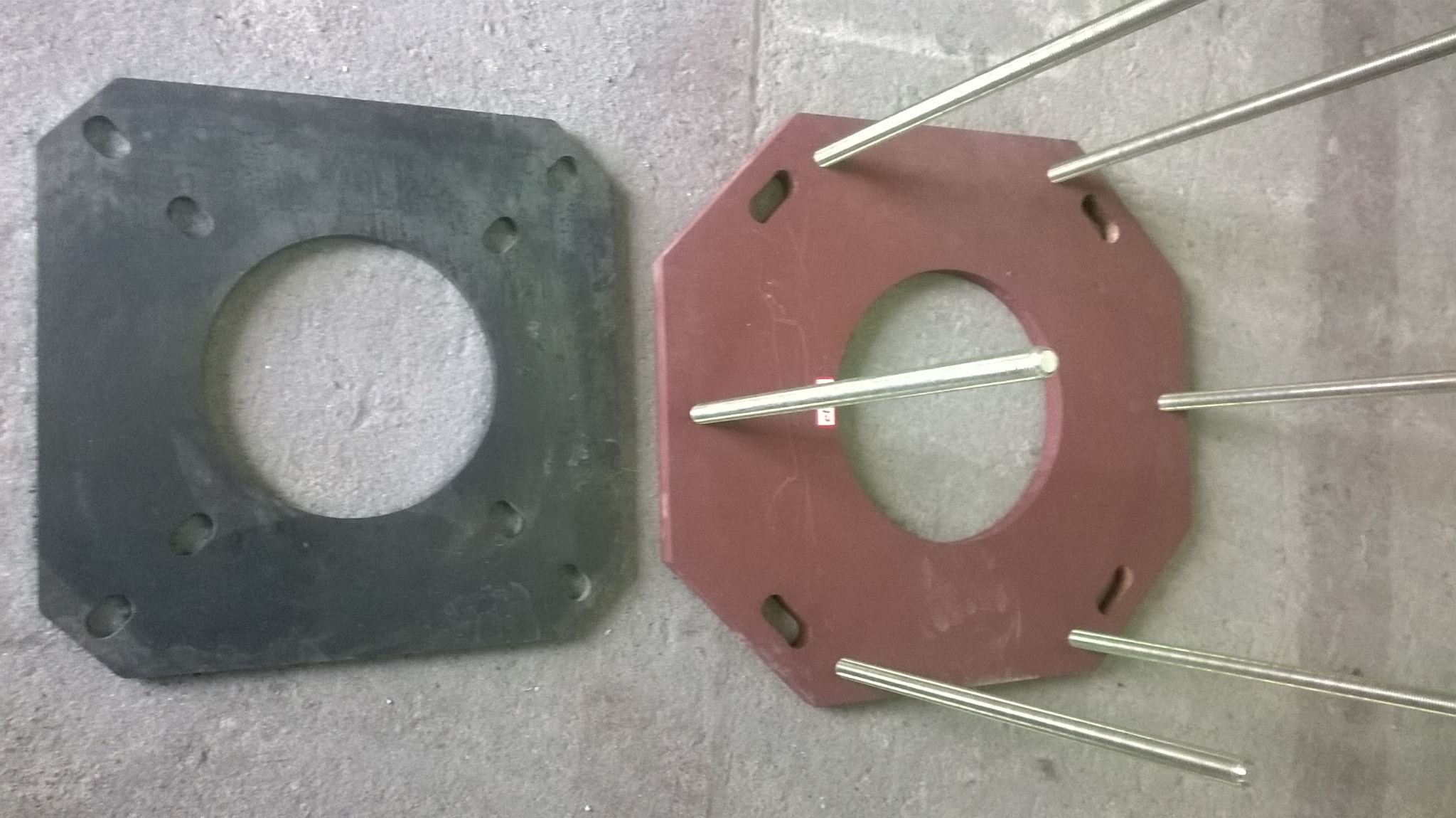 Dwie metalowe płyty z otworami, jedna kwadratowa czarna, druga ośmiokątna czerwona, połączone metalowymi prętami, leżące na szarej betonowej powierzchni.