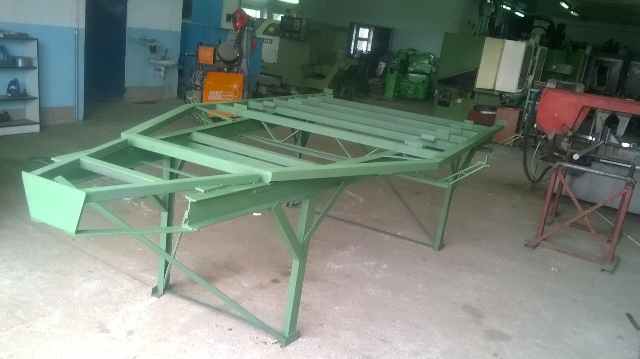 Zielona, metalowa konstrukcja ramy stołu spawalniczego na tle warsztatu z maszynami do obróbki metalu.