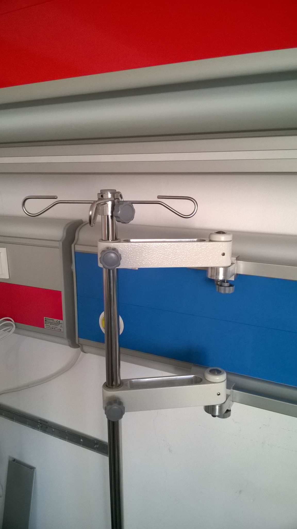 Metalowy, regulowany wieszak na kroplówki przymocowany do ściany w szpitalnej sali, z widocznymi elementami mocującymi i regulacyjnymi.