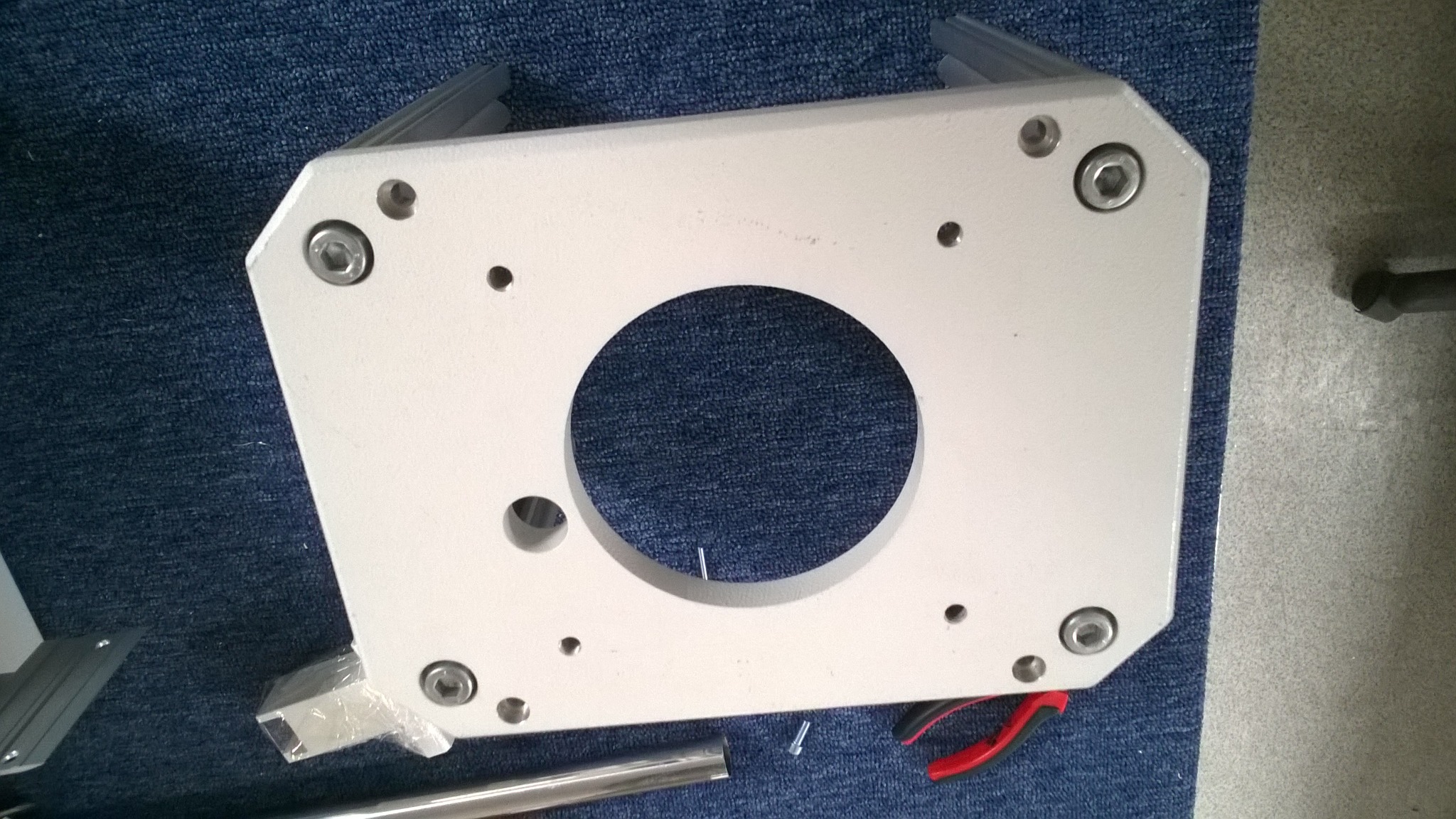 Biała, metalowa płyta z centralnym okrągłym otworem i kilkoma mniejszymi otworami na śruby, leżąca na niebieskiej, teksturowanej powierzchni, obok narzędzi takich jak kombinerki i fragmenty profili...