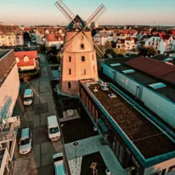 Widok z lotu ptaka na ceglany wiatrak z czterema skrzydłami, otoczony budynkami mieszkalnymi i komercyjnymi, w tle błękitne niebo o zachodzie słońca.