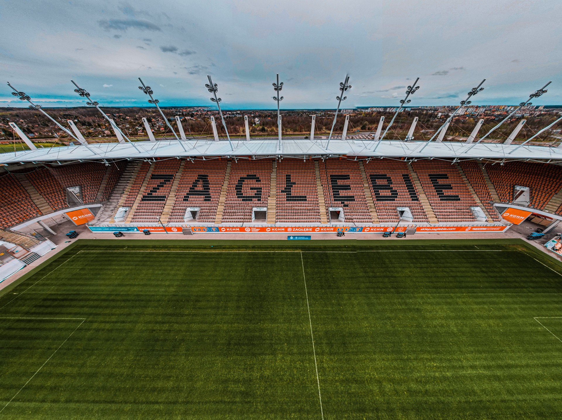 Widok z góry na stadion z napisem 'ZAGŁĘBIE' ułożonym z siedzeń, oświetlony reflektorami, z widocznym miastem w tle.
