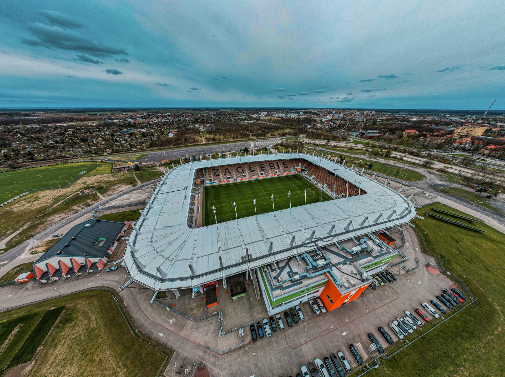 Panoramiczny widok z drona na stadion Zagłębia Lubin z parkingiem, otoczony zielenią i zabudowaniami miejskimi pod pochmurnym niebem.