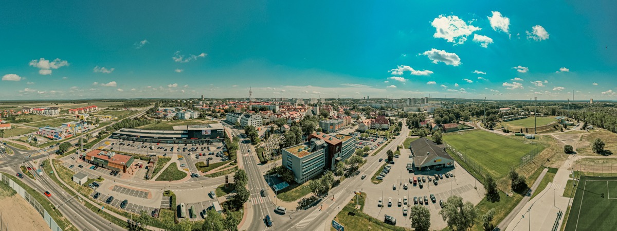 Panoramiczny widok z lotu ptaka na miasto z budynkami mieszkalnymi, centrami handlowymi, stadionem i zielenią w słoneczny dzień z błękitnym niebem i białymi chmurami.
