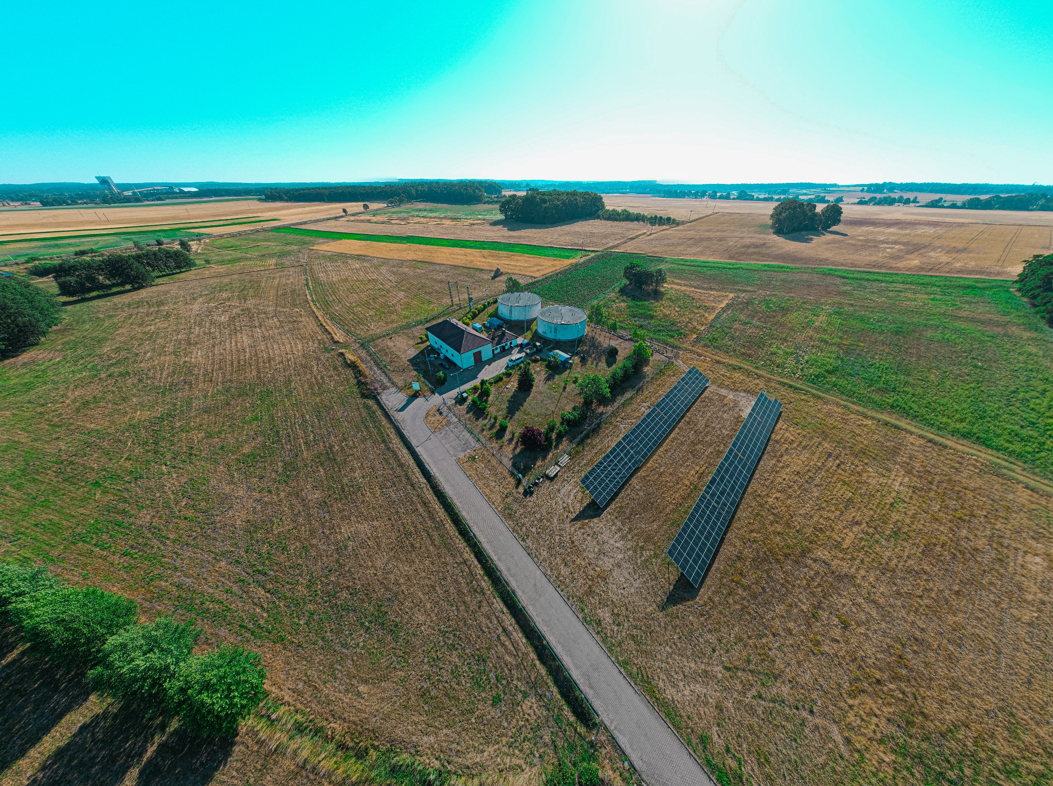 Szeroki kadr z drona przedstawiający farmę fotowoltaiczną z dwoma zbiornikami i budynkiem technicznym pośród pól uprawnych, z widocznym w oddali lasem i konstrukcją przemysłową.