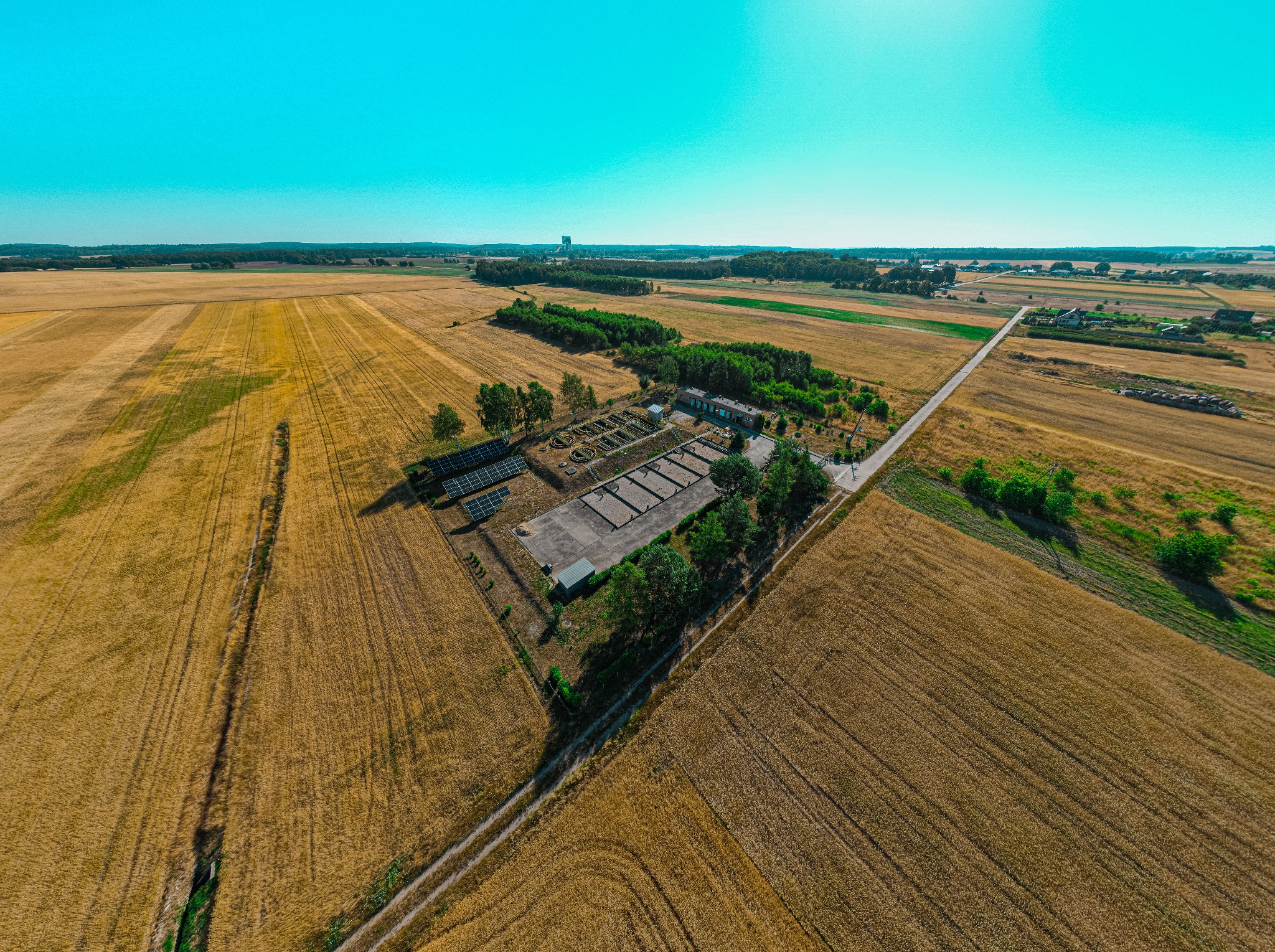 Szeroki kadr z lotu ptaka ukazuje pole po żniwach, drogę polną, niewielki kompleks budynków z oczyszczalnią ścieków i panelami słonecznymi, otoczony zielenią drzew i krzewów, z lasem...