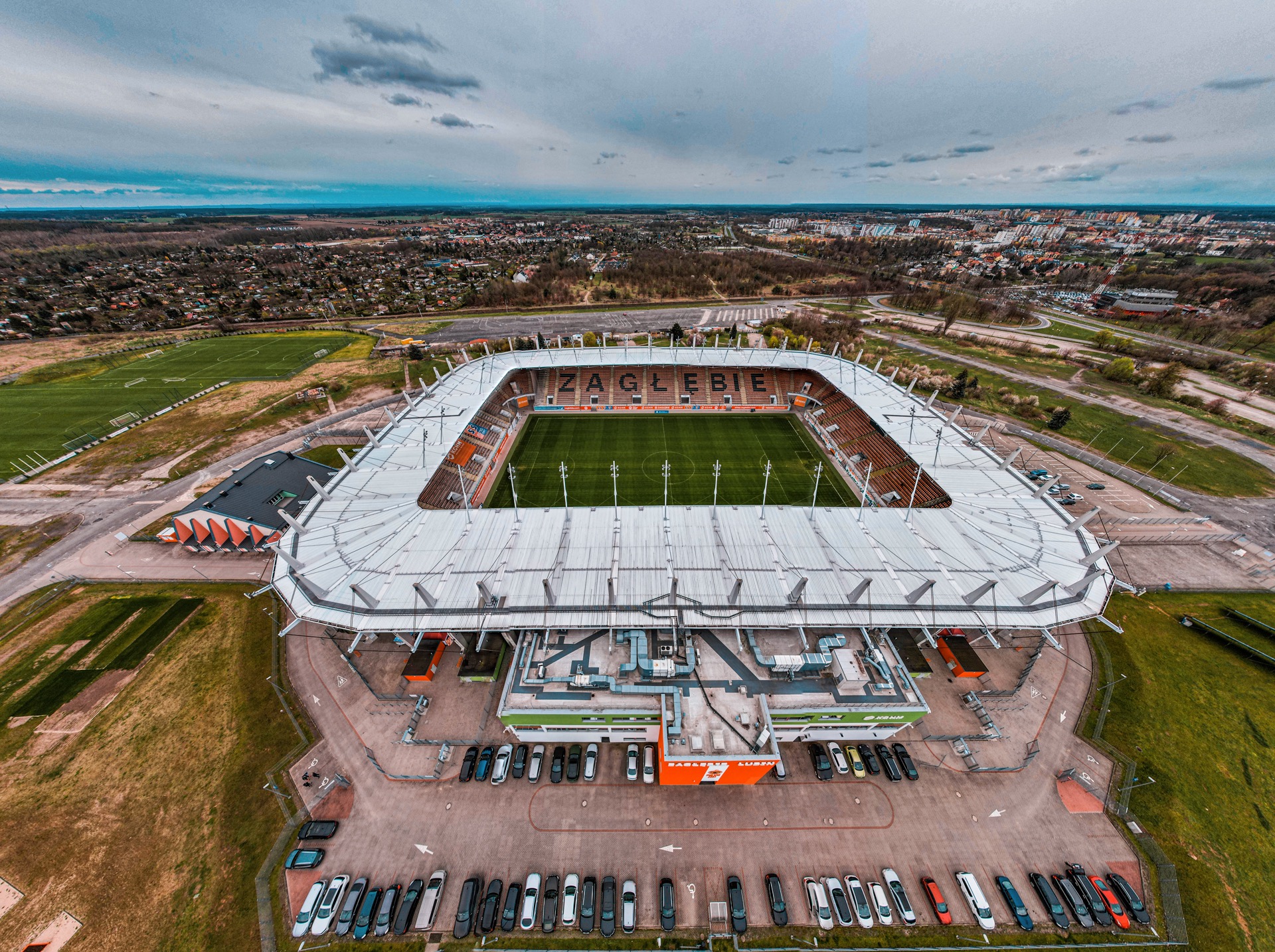 Widok z drona na stadion piłkarski Zagłębia Lubin, z parkingiem pełnym samochodów i miastem w tle pod pochmurnym niebem.