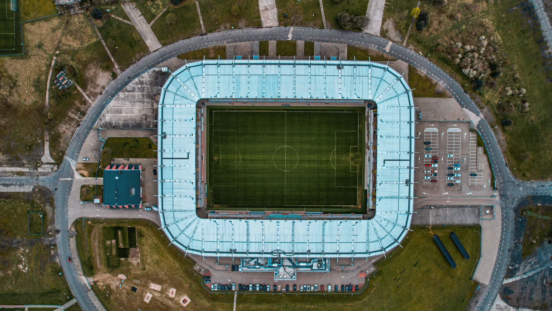 Widok z drona na stadion piłkarski z parkingiem i drogami dojazdowymi, otoczony zielenią.