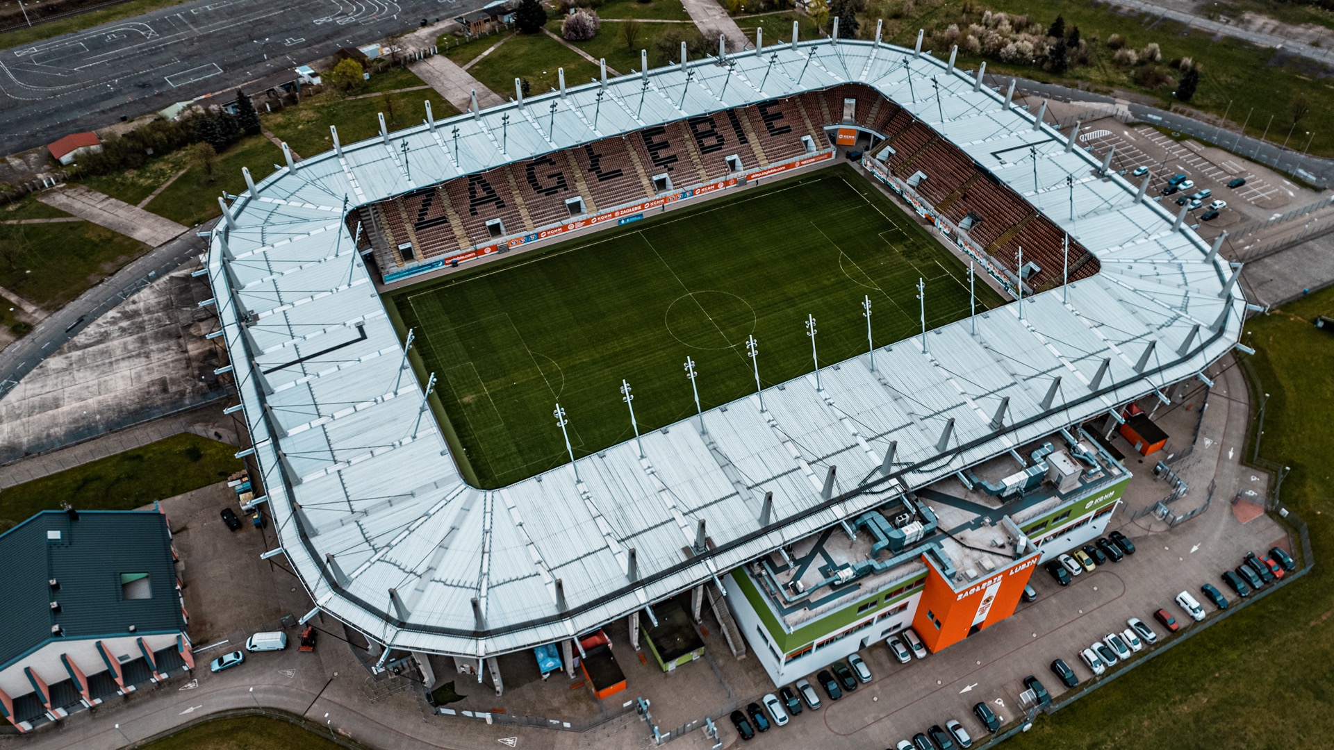 Widok z lotu ptaka na stadion Zagłębia Lubin z zaparkowanymi samochodami i otaczającym terenem.