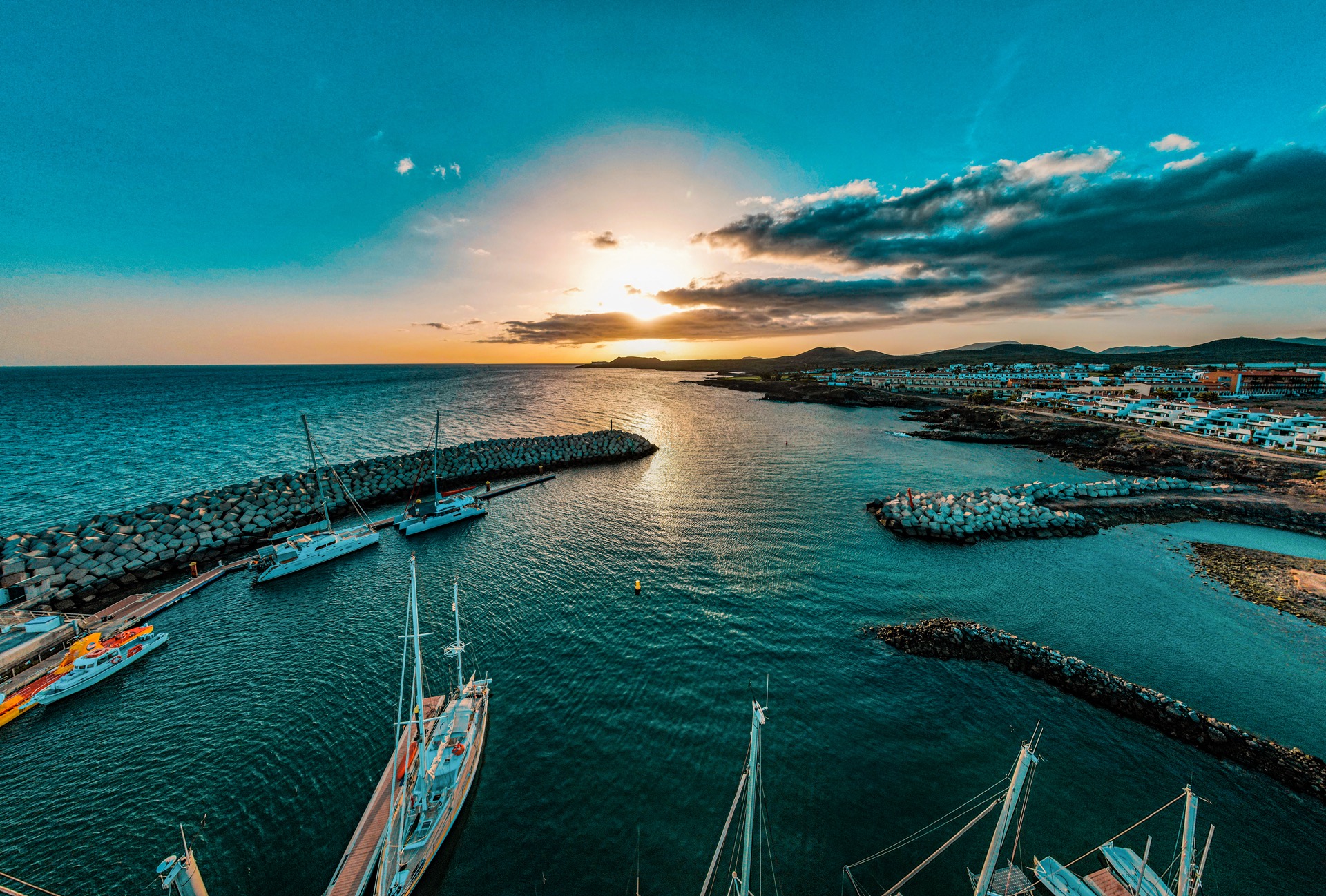 Szeroki kadr na port jachtowy o zachodzie słońca, falochrony z bloków skalnych, budynki na brzegu w oddali, jachty cumujące przy nabrzeżu.