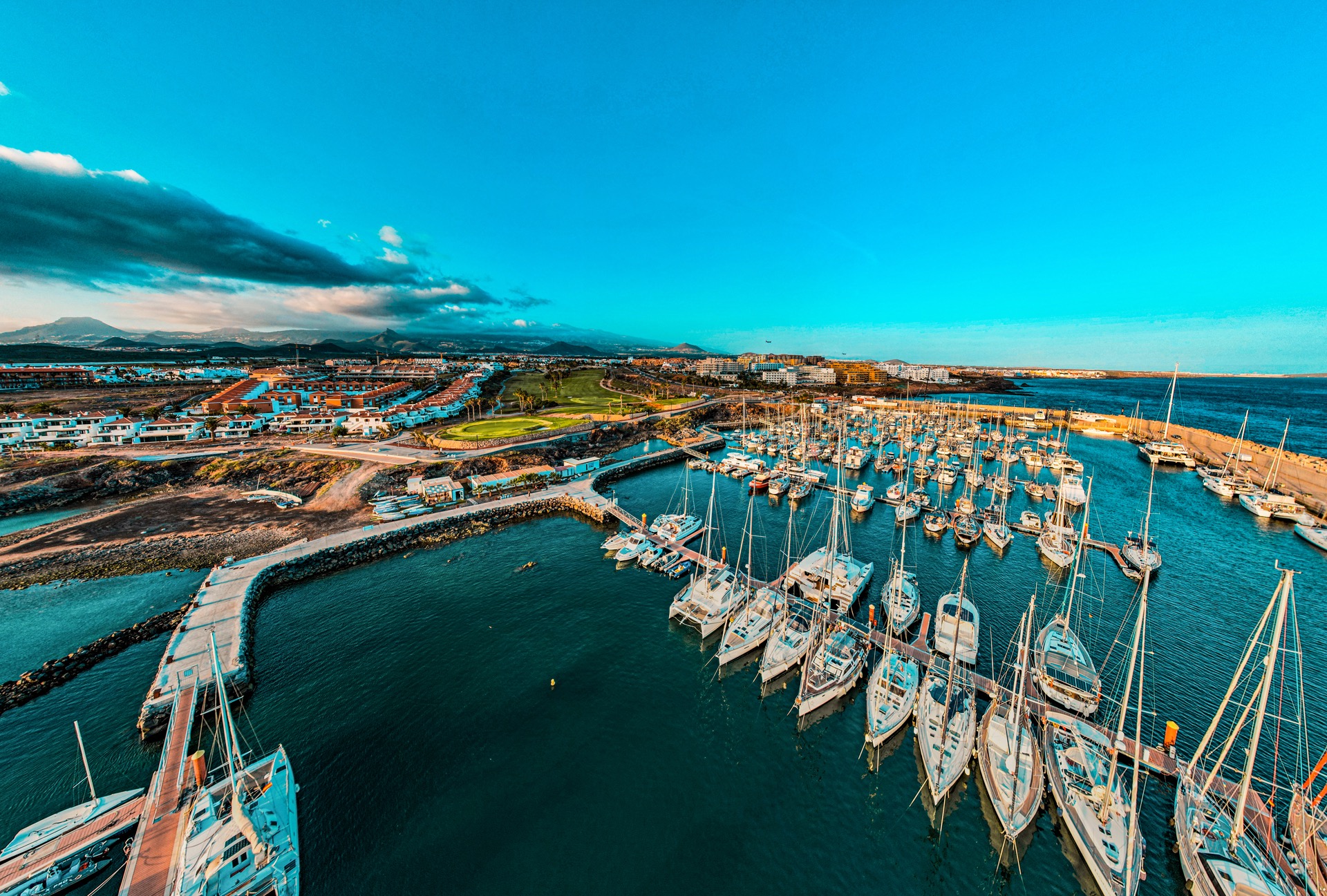 Panoramiczny widok z lotu ptaka na port jachtowy z licznymi żaglówkami i katamaranami, w tle zabudowa miejska, pole golfowe i góry pod błękitnym niebem.