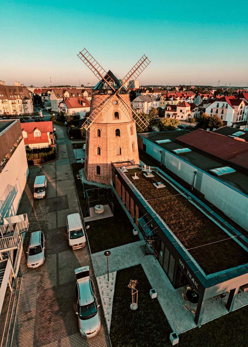 Widok z lotu ptaka na ceglany wiatrak z czterema skrzydłami, otoczony budynkami mieszkalnymi i komercyjnymi, w tle błękitne niebo o zachodzie słońca.
