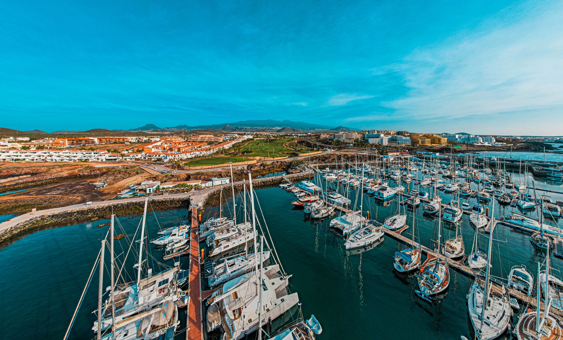 Panoramiczne ujęcie portu jachtowego z licznymi żaglówkami zacumowanymi przy pomostach, w tle zabudowa miejska z czerwonymi dachami i zielone pole golfowe, a w oddali zarys gór pod błękitnym niebem.