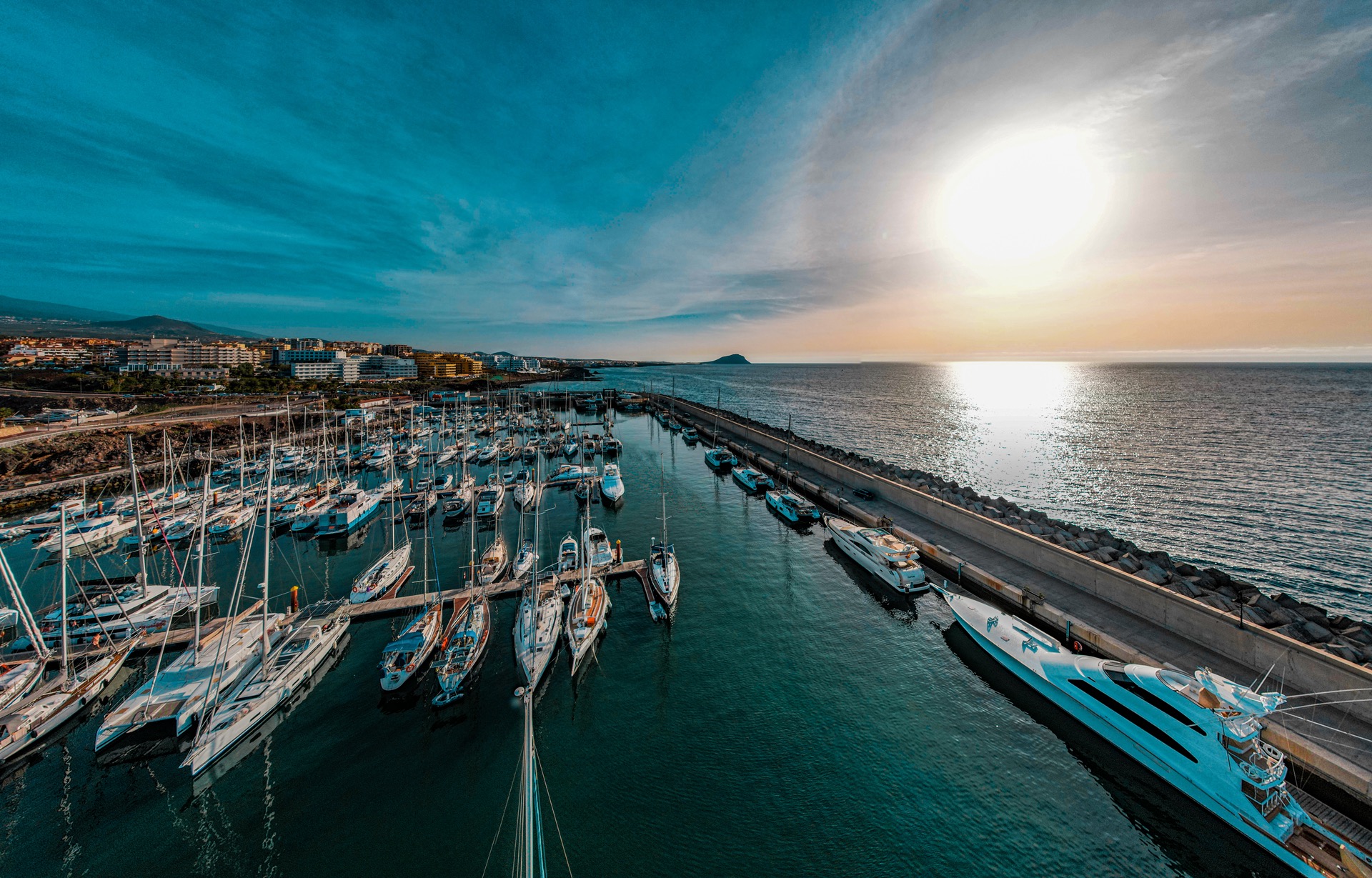 Panorama portu jachtowego o zachodzie słońca. Widok z lotu ptaka na zacumowane jachty, falochron i budynki w tle. Intensywne kolory nieba i odbicia w wodzie.