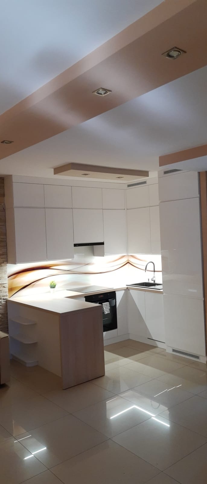 Nowoczesna, biała kuchnia z barkiem w Wodzisławiu Śląskim, z dekoracyjnym panelem szklanym nad blatem i subtelnym oświetleniem.