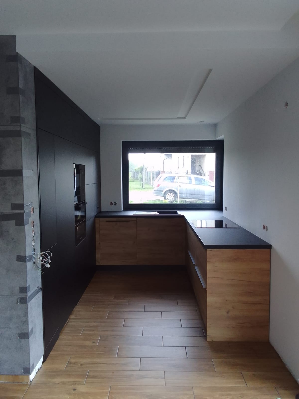 Nowoczesna kuchnia z drewnianymi frontami i czarnym blatem, widok na okno z roletą, fragment ściany w trakcie wykończenia z widocznymi przewodami elektrycznymi.