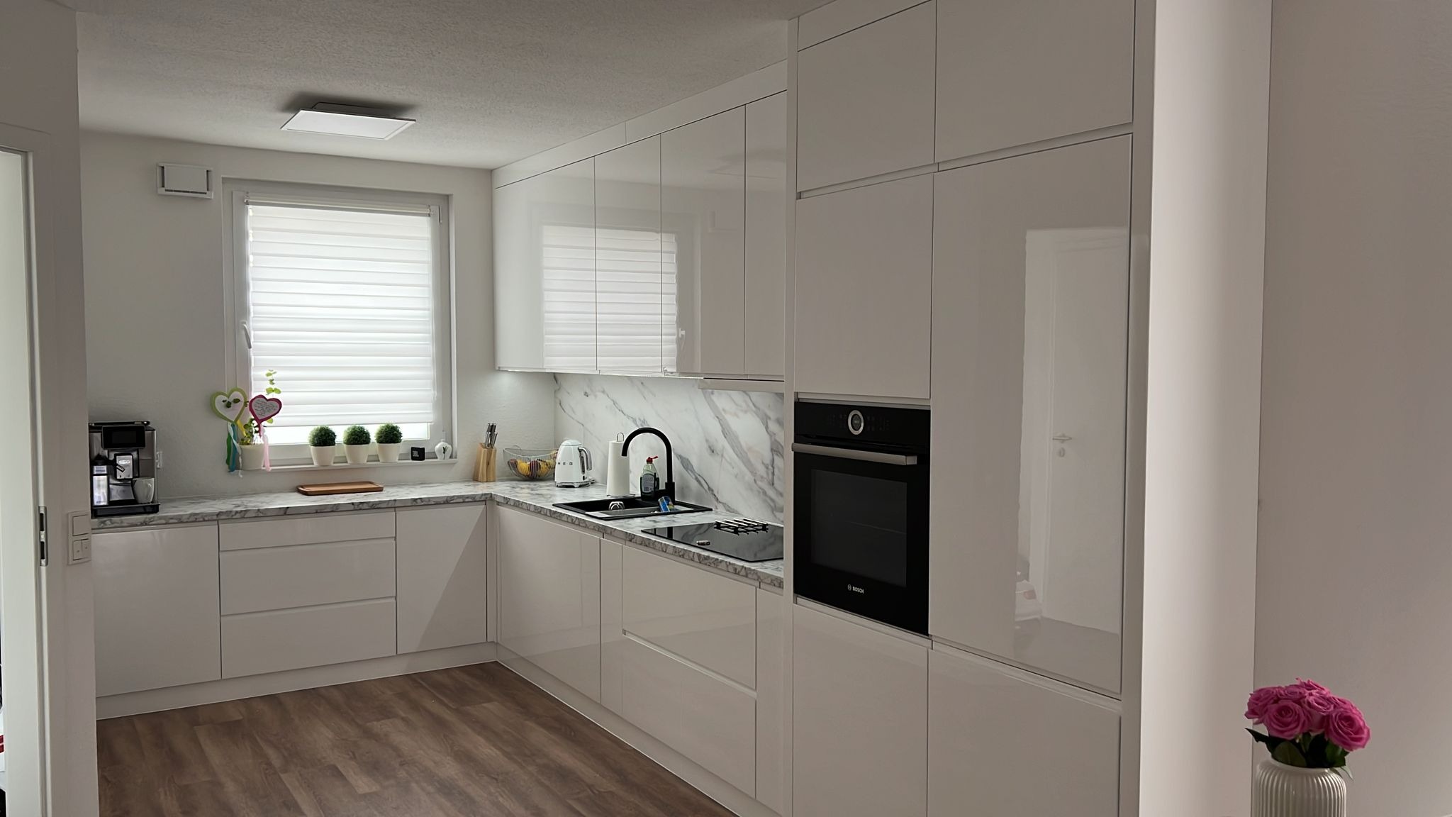 Nowoczesna, jasna kuchnia z białymi, lakierowanymi frontami, marmurowym blatem i backsplashiem, czarną armaturą i wbudowanym piekarnikiem. Na blacie przy oknie ekspres do kawy i dekoracje.