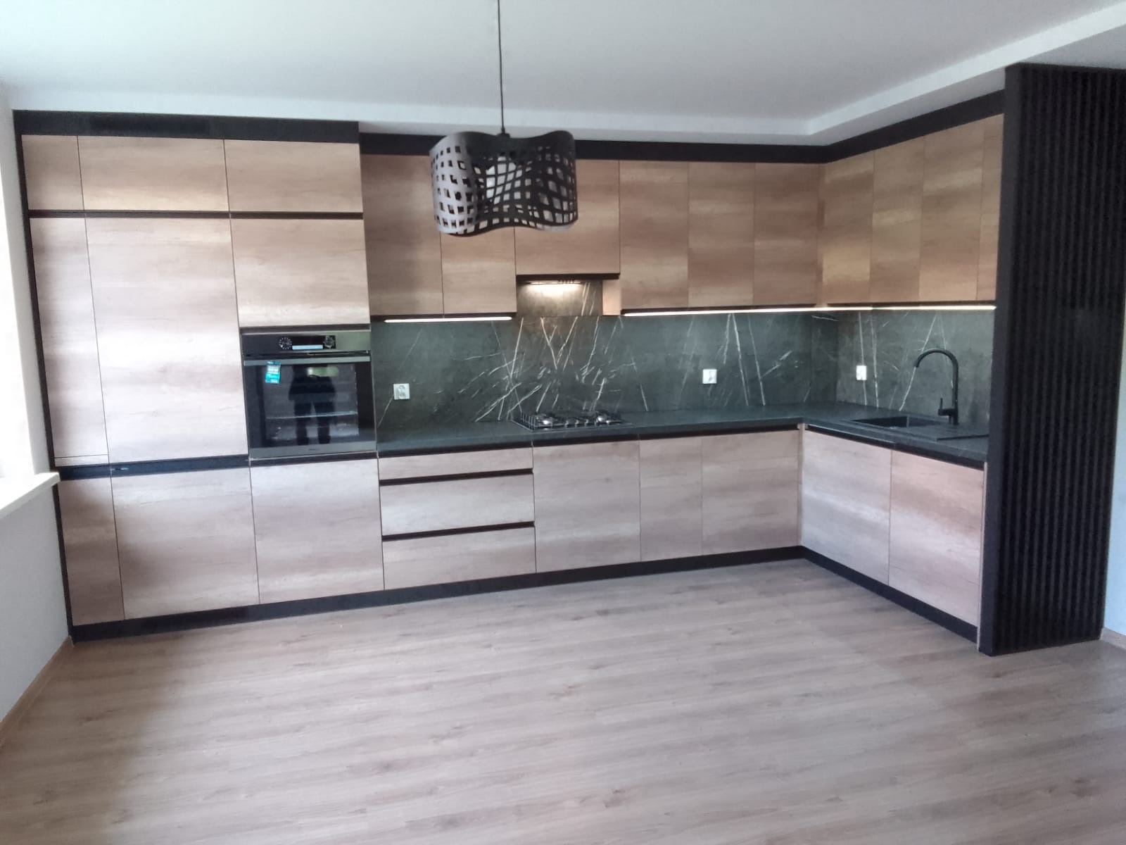 Jasna, nowoczesna kuchnia z drewnopodobnymi frontami, czarnymi akcentami i marmurkowym backsplashiem, oświetlona listwą LED pod szafkami wiszącymi i designerską lampą wiszącą.