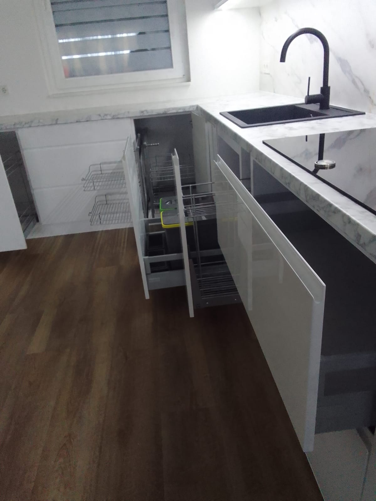 Nowoczesna kuchnia z białymi, błyszczącymi frontami, marmurowym blatem i czarną armaturą. Widoczne wysuwane kosze cargo w szafce narożnej i szafka z metalowymi koszami do przechowywania.