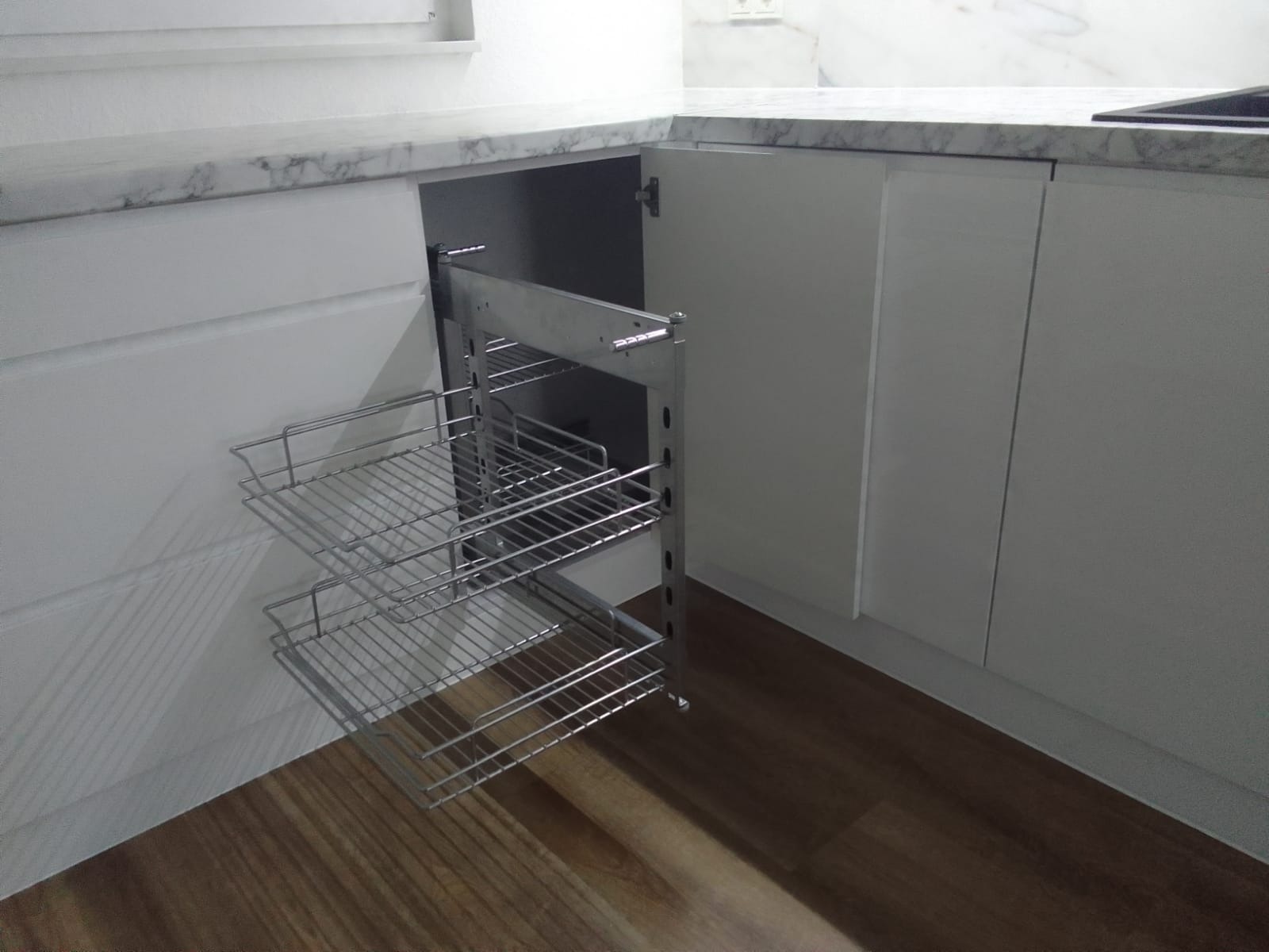 Wysuwany, dwupoziomowy kosz cargo zamontowany w narożnej szafce kuchennej z białymi frontami i marmurowym blatem, widoczna drewniana podłoga.