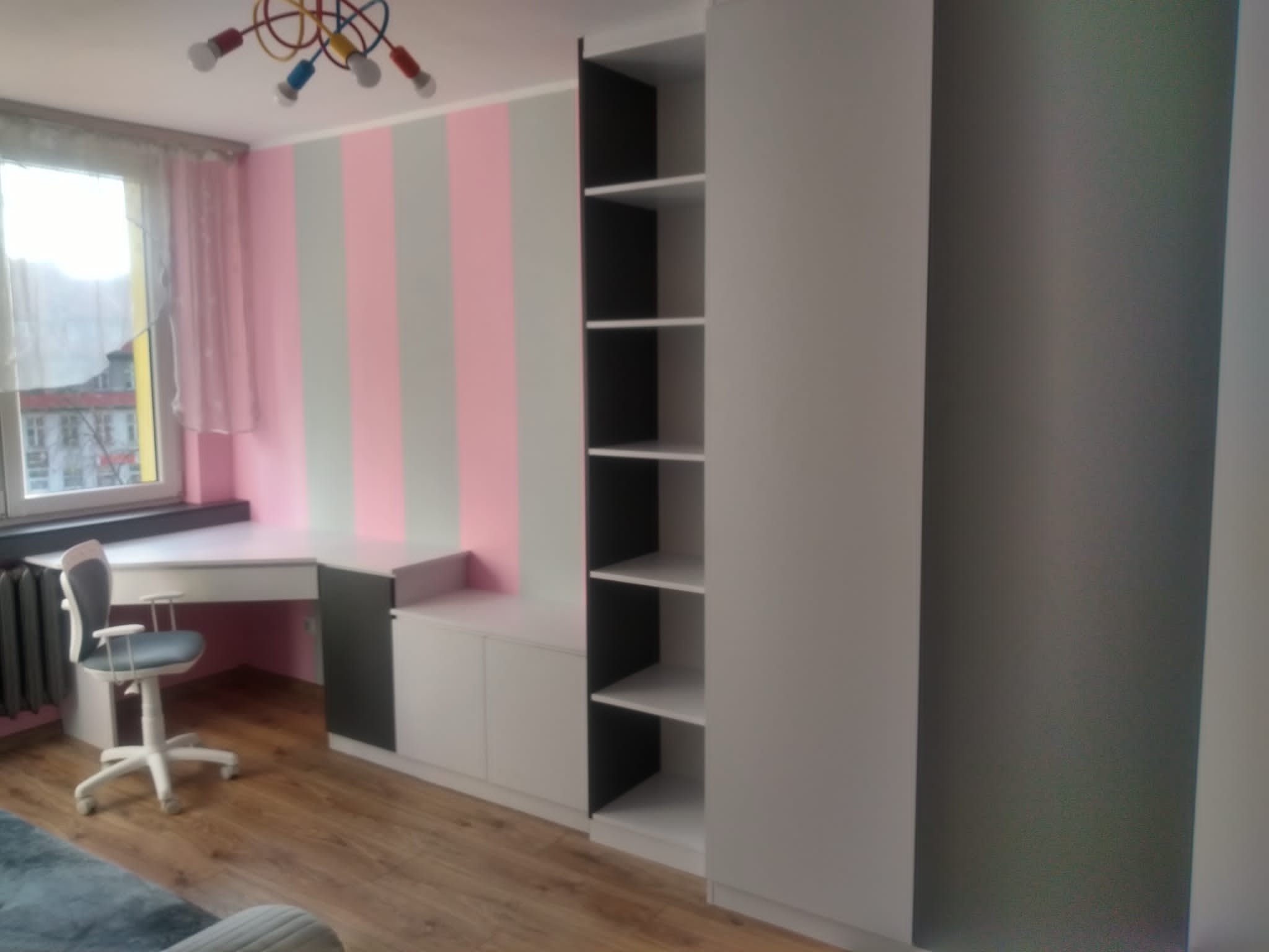 Pokój dziecięcy z zabudową meblową: biurko narożne z szarą tapicerowaną krzesłem na kółkach, szafki i regał z półkami na tle ściany w różowo-szare pasy, podłoga z paneli.