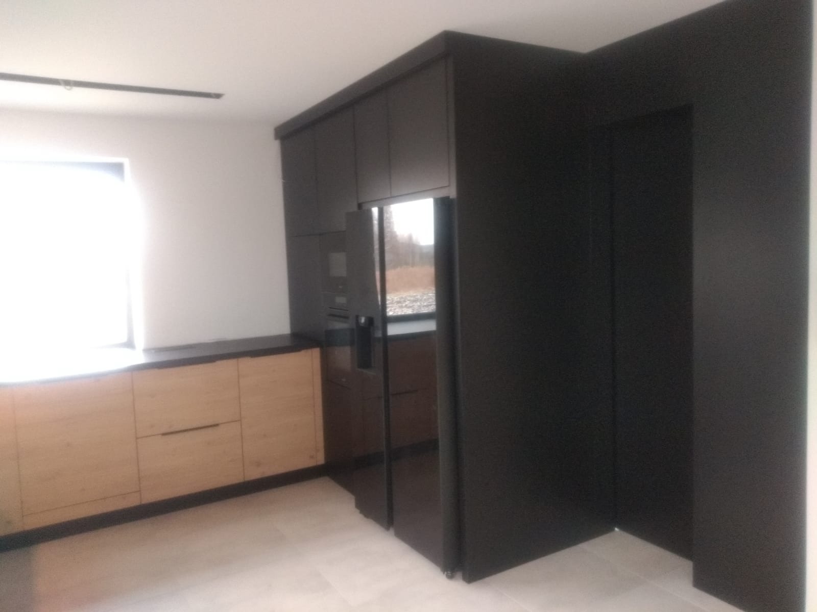 Nowoczesna kuchnia z drewnianymi frontami szafek dolnych i czarnymi szafkami górnymi zabudowanymi do sufitu, zintegrowana z czarną ścianą i lodówką.