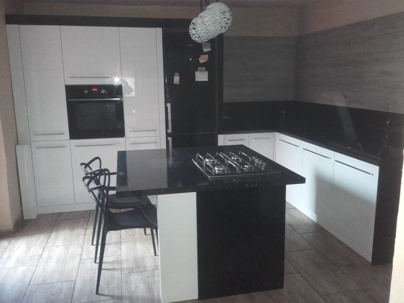 Nowoczesna kuchnia z białymi szafkami, czarnym blatem i wyspą z płytą grzewczą, oświetlona wiszącą lampą w stylu origami.