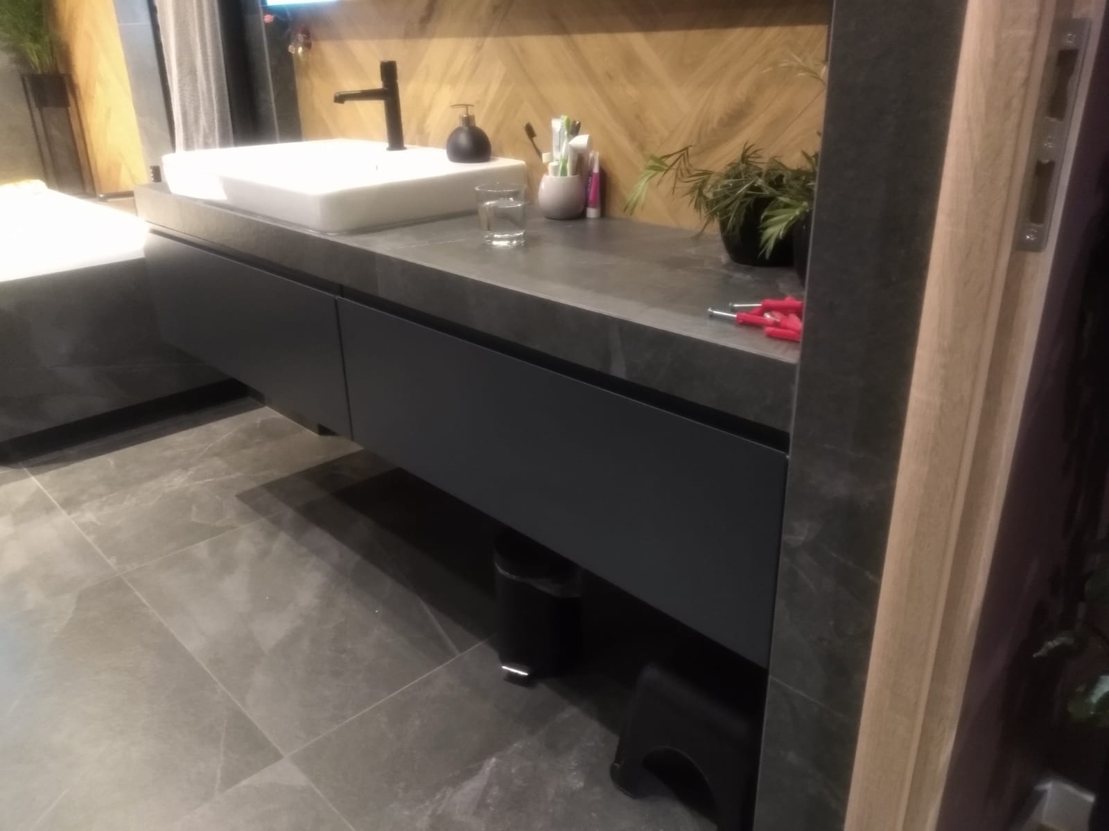 Szara szafka pod umywalkę z blatem z kamienia, nowoczesna bateria, drewniana ściana w jodełkę, szare płytki podłogowe, akcesoria łazienkowe i narzędzia na blacie.