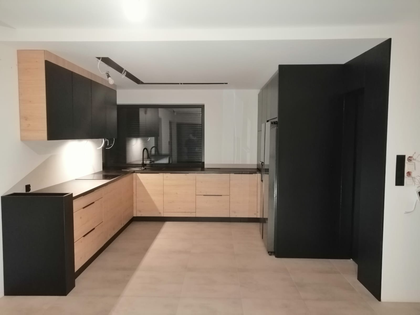 Nowoczesna kuchnia w odcieniach drewna i czerni, z widocznym oświetleniem podszafkowym i minimalistyczną armaturą, uchwycona z szerokiego kąta w jasnym wnętrzu.