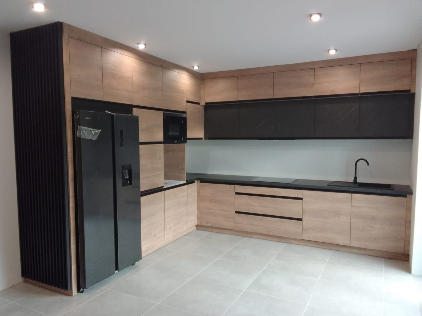 Nowoczesna kuchnia z zabudową w odcieniach drewna i czerni, z dużą lodówką side-by-side i czarnym blatem, oświetlona punktowymi lampami sufitowymi.