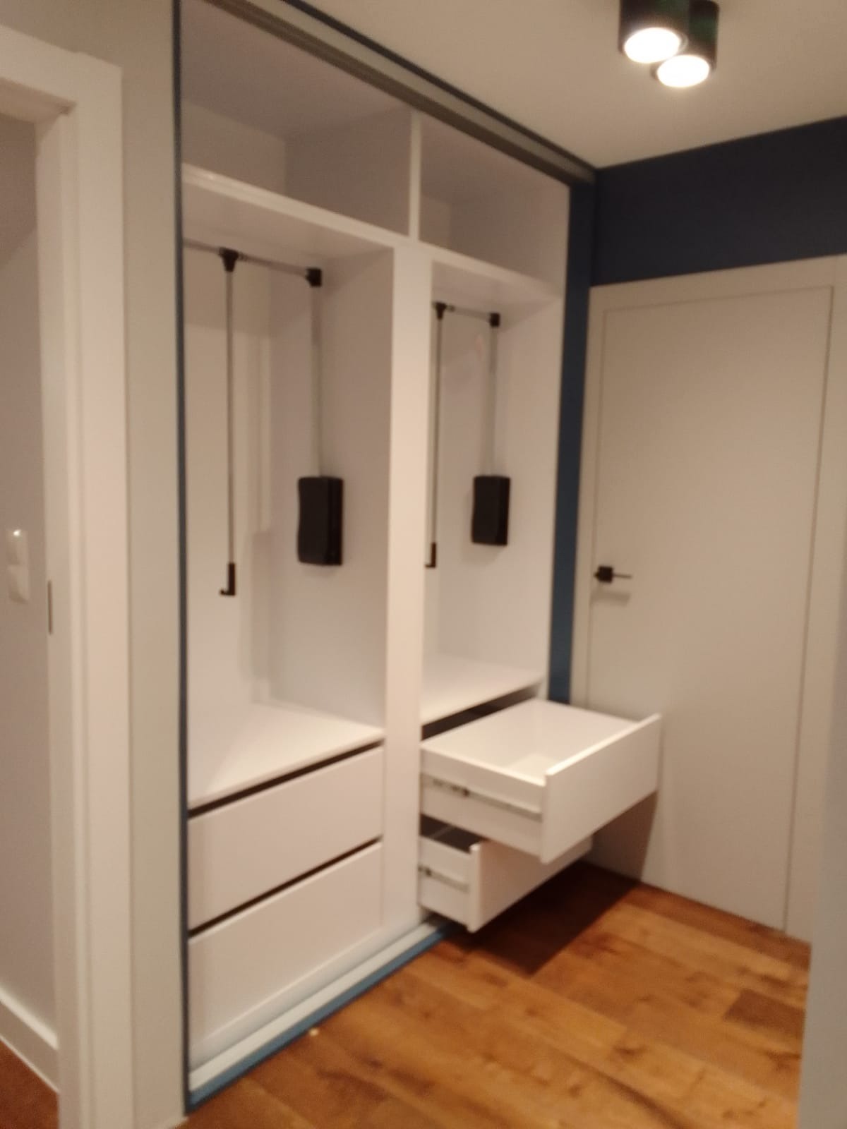 Biała szafa wnękowa z wysuwanymi szufladami i drążkami na ubrania, widok z boku na drewnianą podłogę.