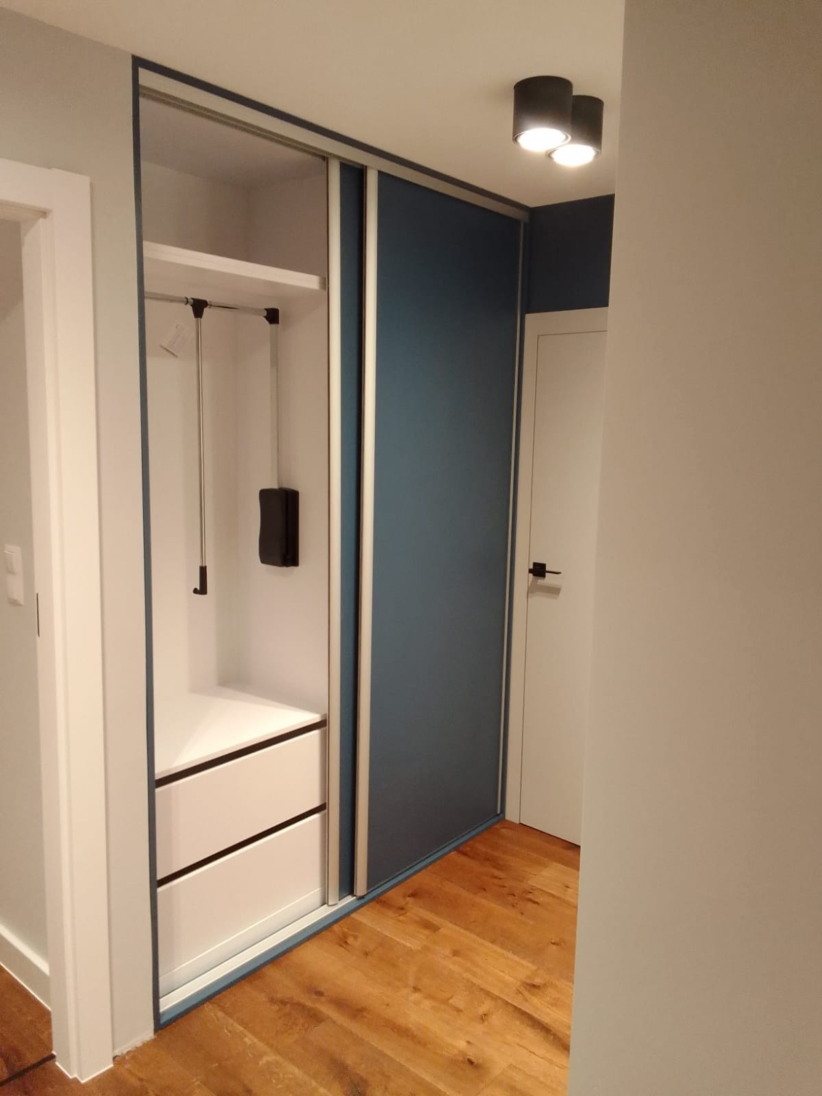 Wnętrze przedpokoju z szafą wnękową z przesuwnymi drzwiami w kolorze granatowym, z widocznym systemem szuflad i drążkiem na ubrania, oświetlonymi przez sufitowe lampy.