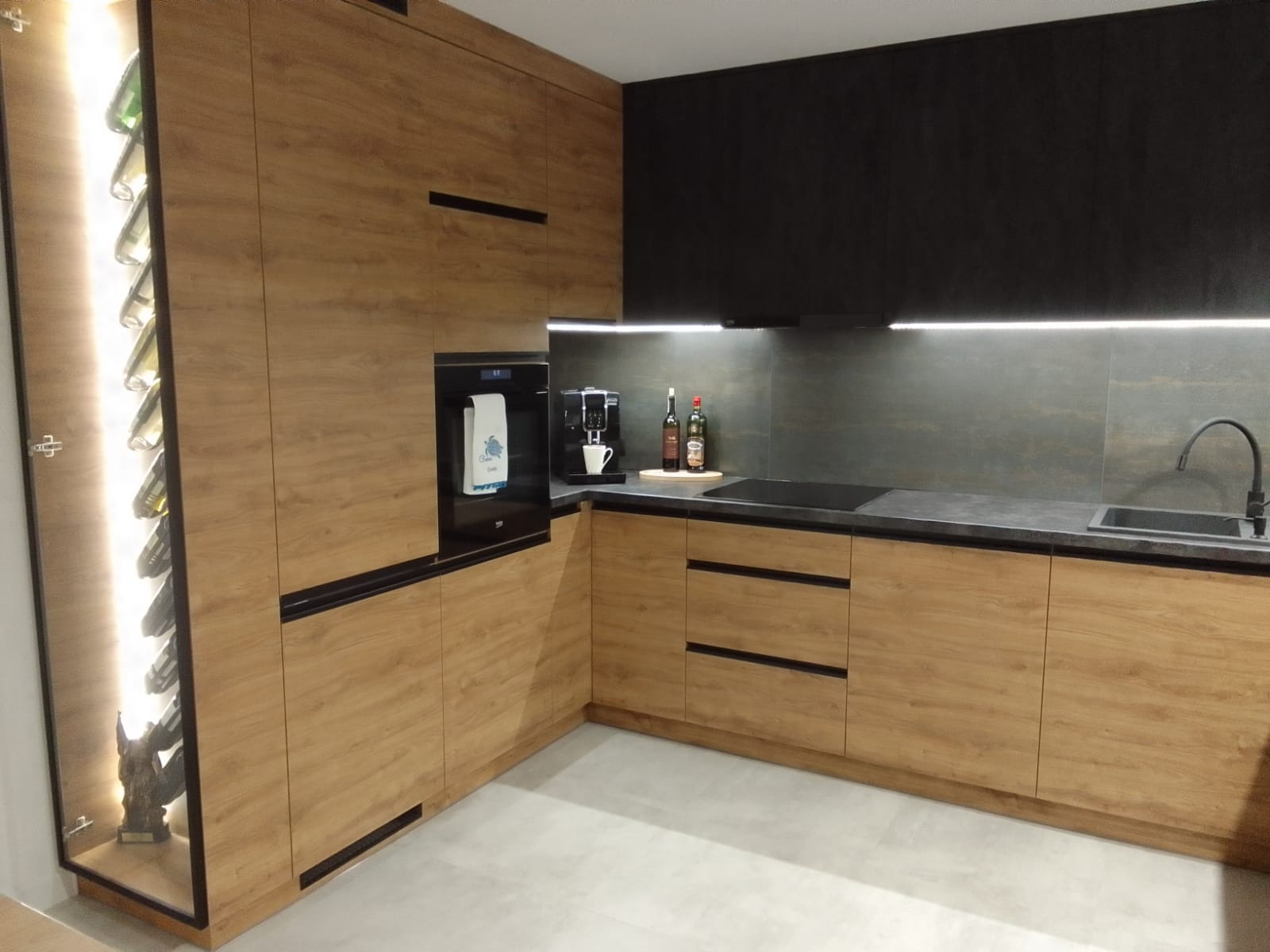 Nowoczesna kuchnia z drewnianymi frontami, czarnym blatem i ścianą, oświetleniem LED pod szafkami wiszącymi i w zabudowanej wnęce na wino z dekoracją.